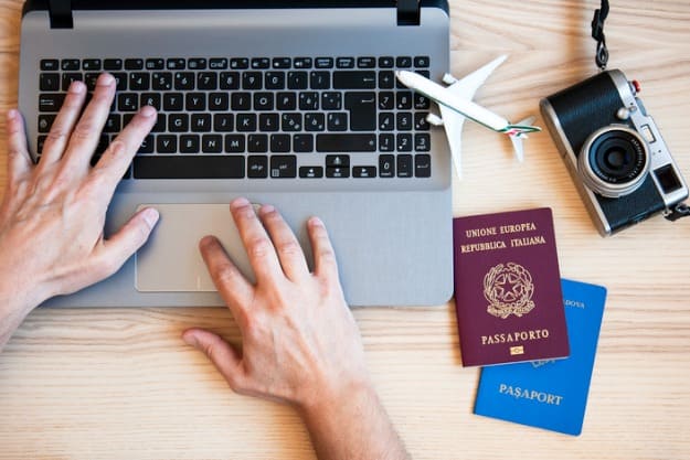 Mãos de homem digitando em notebook com câmera, passaporte e avião de brinquedo ao lado.