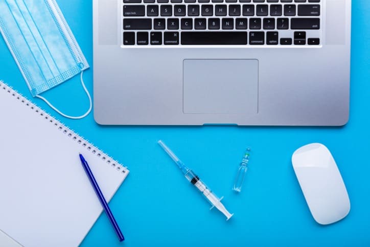 Notebook, seringa e caderno em cima da mesa, representando a tradução juramentada da carteira de vacinação.