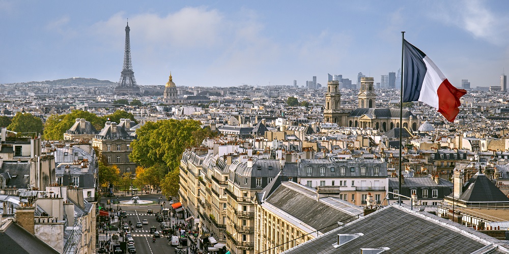 Vista panorâmica de Paris, contendo a bandeira da França em primeiro plano e a Torre Eiffel de fundo